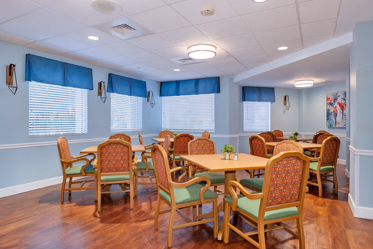 A bright dining room with multiple wooden tables and chairs featuring green cushions and patterned backs. The room has light blue walls, three windows with blue valances, and wooden flooring. Small potted plants are placed on the tables, and there is a colorful painting on one wall.