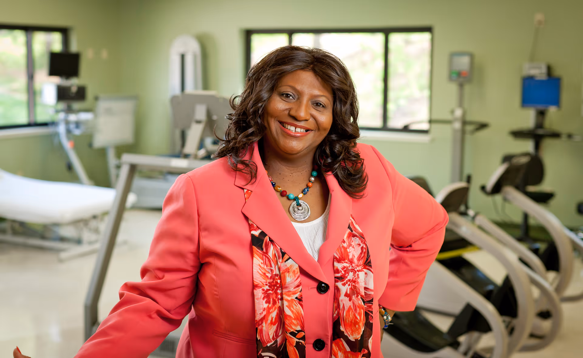 A smiling woman wearing a bright pink blazer and floral scarf stands in a rehabilitation or physical therapy room with exercise equipment and medical devices in the background.