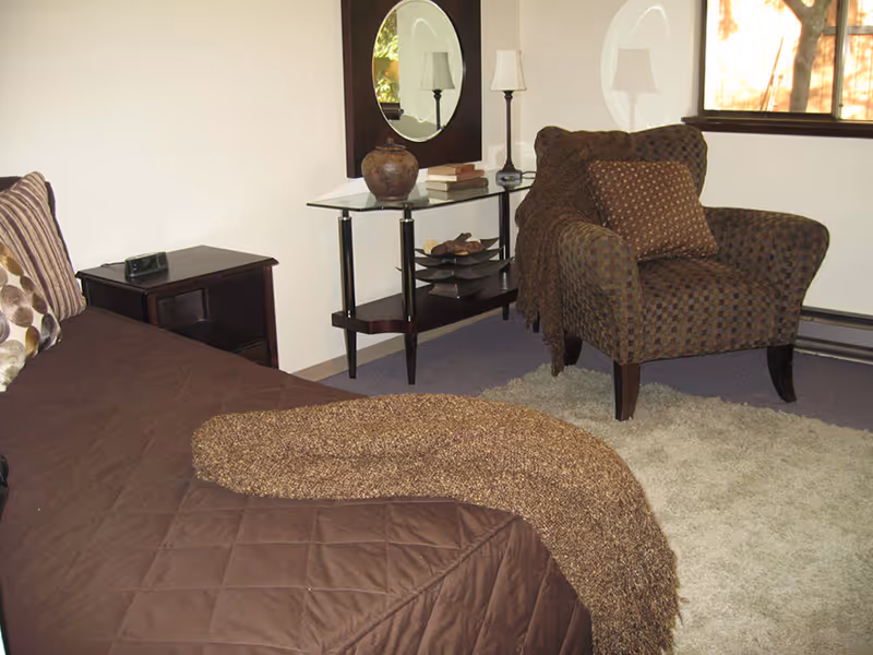 Cozy bedroom with a brown quilted bed, patterned armchair, glass console table, and area rug.