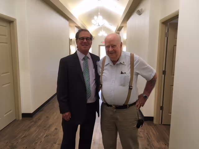 Two elderly men standing in a well-lit hallway with wooden flooring and cream-colored walls. One man is wearing a dark suit with a green tie and glasses, and the other man is wearing a white short-sleeve shirt with suspenders and beige pants. Both are smiling and posing for the photo.
