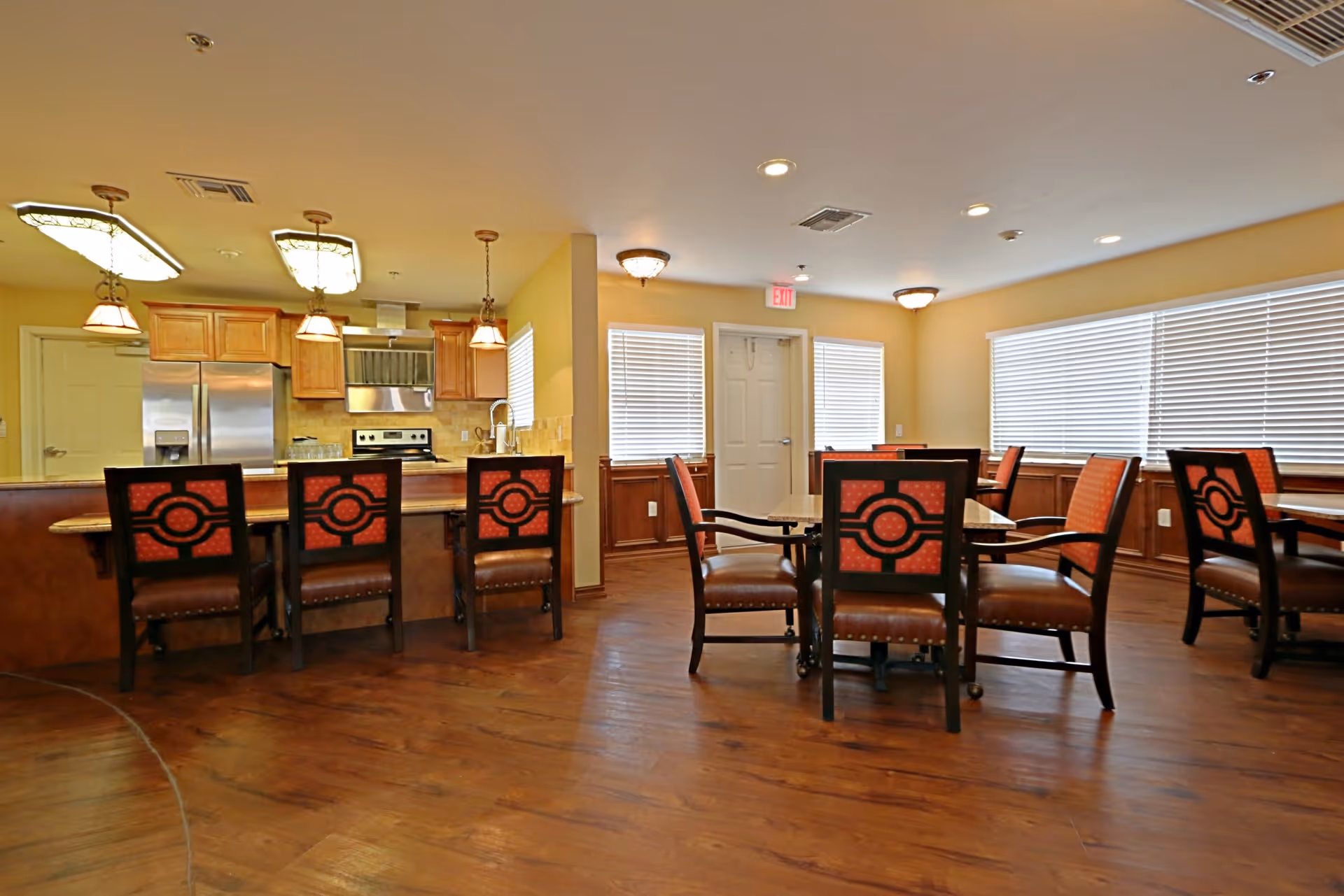 Interior view of a senior living facility dining area with wooden floors, multiple tables and chairs with red cushioned seats, and a kitchen area with stainless steel appliances and pendant lighting in the background.