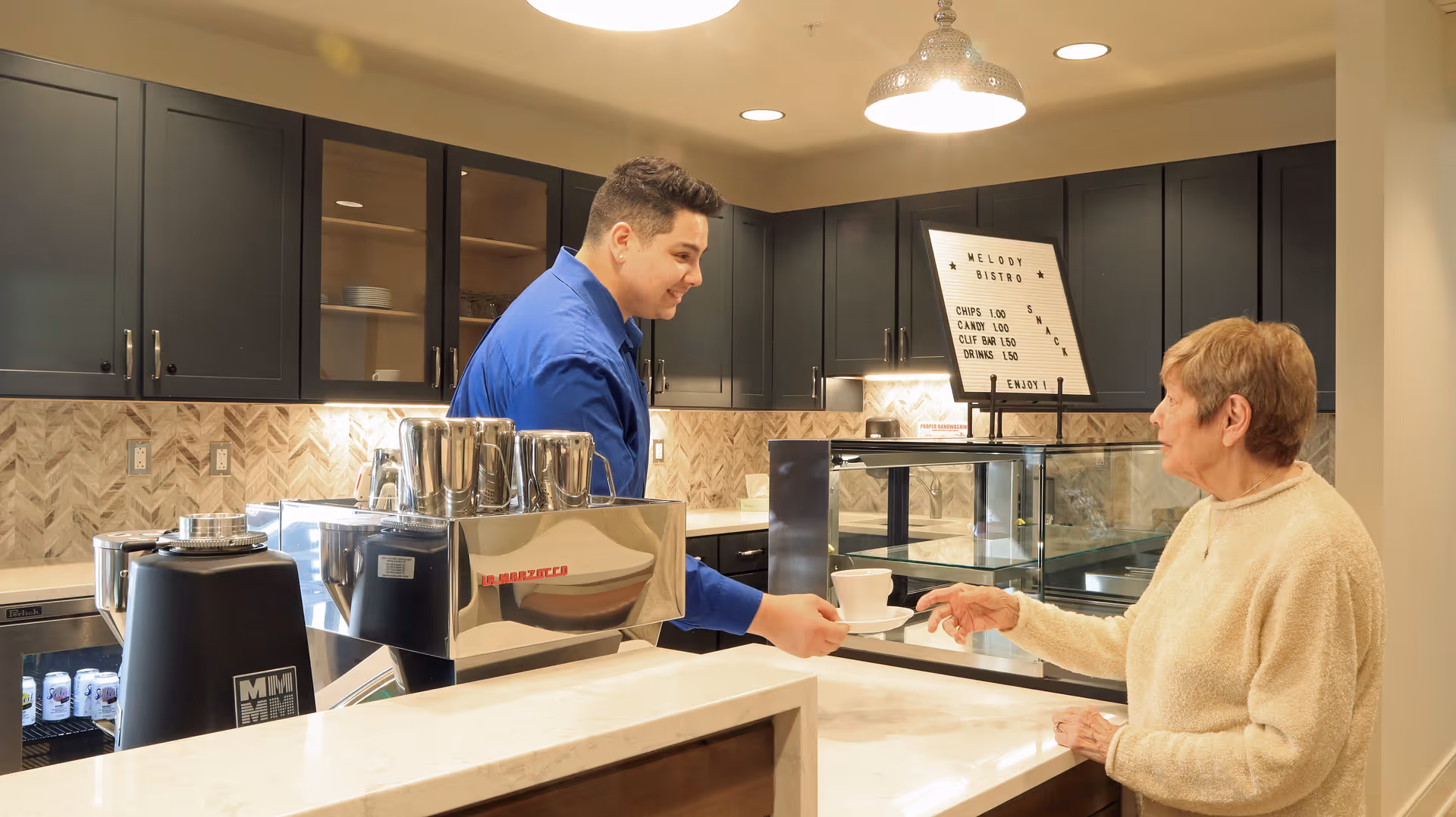 A young man in a blue shirt serves a cup of coffee to an elderly woman wearing a beige sweater at a counter in a modern kitchen or bistro area with dark cabinets and a light-colored countertop.
