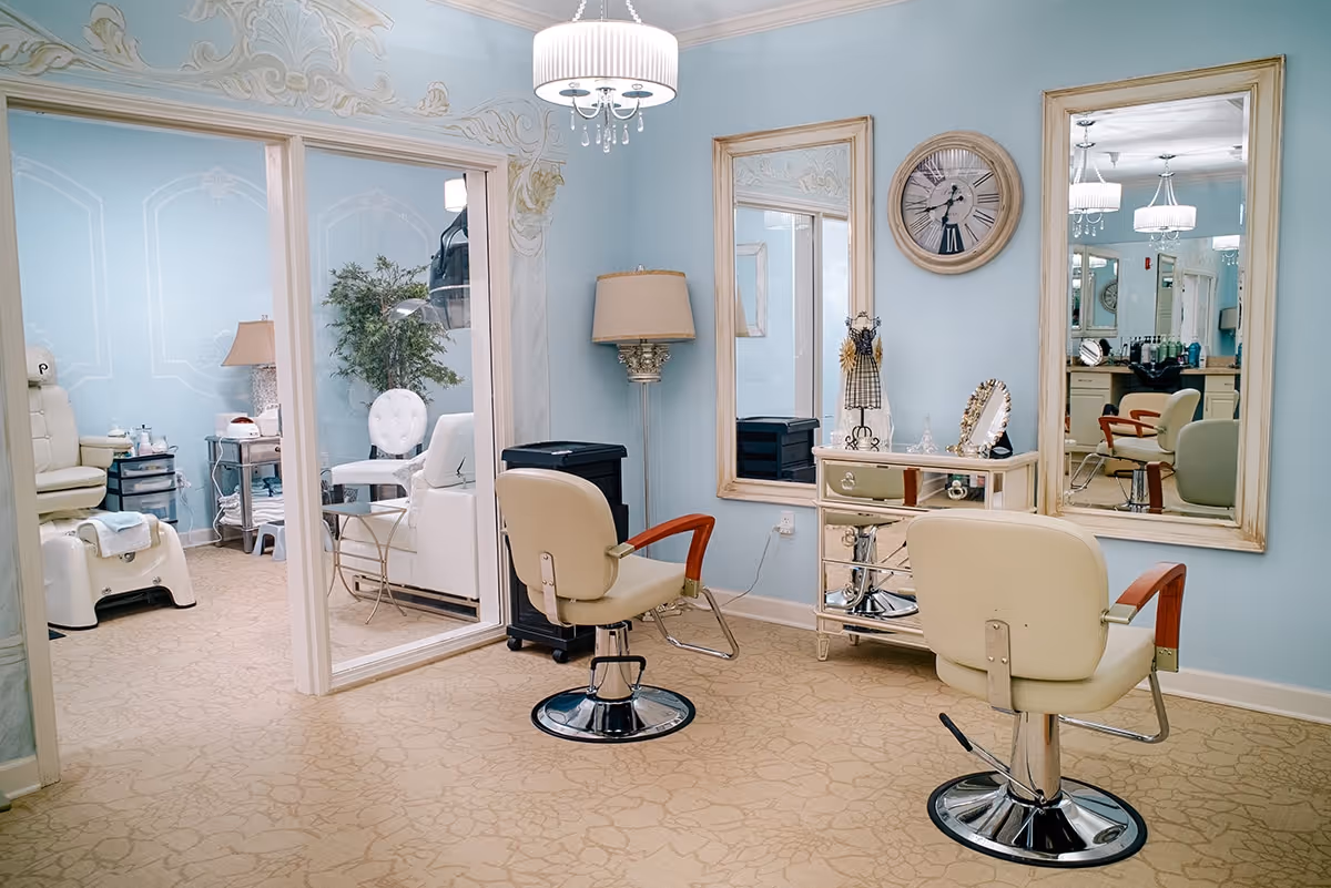 Interior view of a salon area in a senior living facility with two beige salon chairs facing large mirrors on a light blue wall. The room features a decorative wall clock, a lamp, and a mirrored cabinet with small decorative items. Through a glass partition, a pedicure station with a white chair and foot bath is visible in an adjoining room.