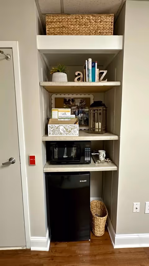 A built-in shelving unit with a woven basket on the top shelf, decorative letters and books on the second shelf, a framed photo, a lantern, and a tissue box on the third shelf, a microwave and a cup on a tray on the fourth shelf, and a black mini refrigerator with a woven basket on the floor next to it. The shelving unit is set against a beige wall with a door to the left and electrical outlets to the right.