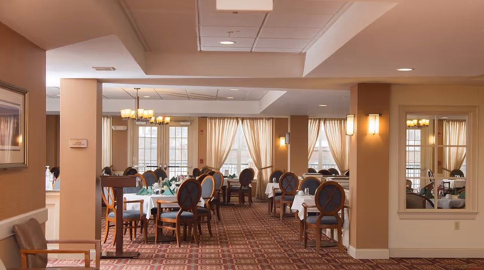 Bright dining room with tables set with white tablecloths and blue-upholstered chairs, chandeliers, and large windows with curtains.