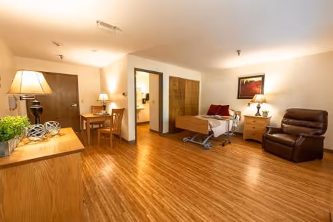 A spacious senior living room with wooden flooring featuring a hospital-style bed with red pillows, a brown recliner chair, a wooden nightstand with a lamp and framed artwork above it, a wooden table with two chairs, and a wooden cabinet with decorative items and a lamp.