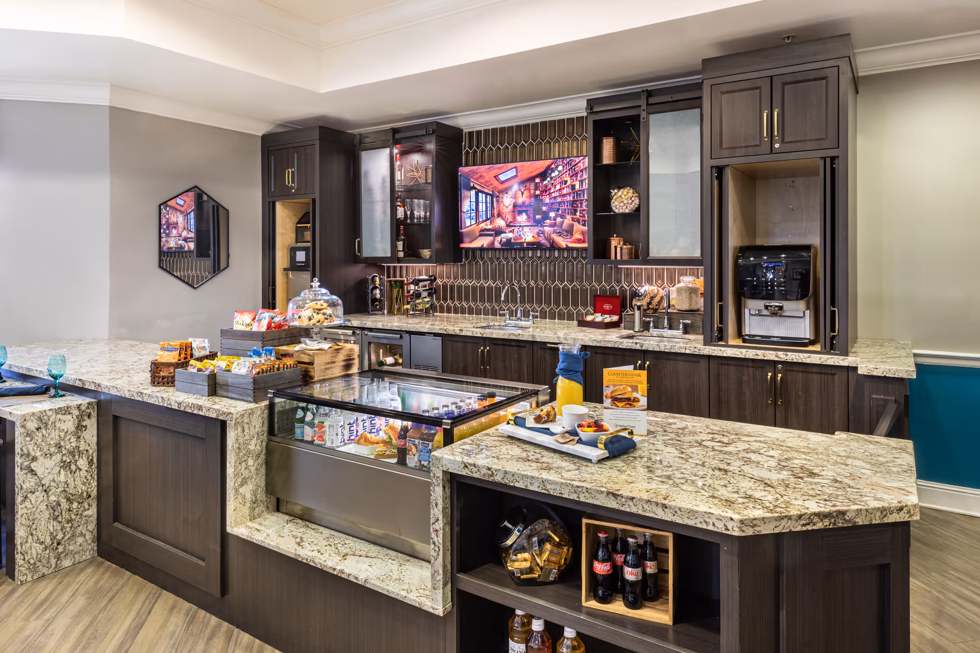 Modern communal kitchenette/snack bar with granite countertops, dark wood cabinets, a beverage machine, display cooler and assorted snacks.