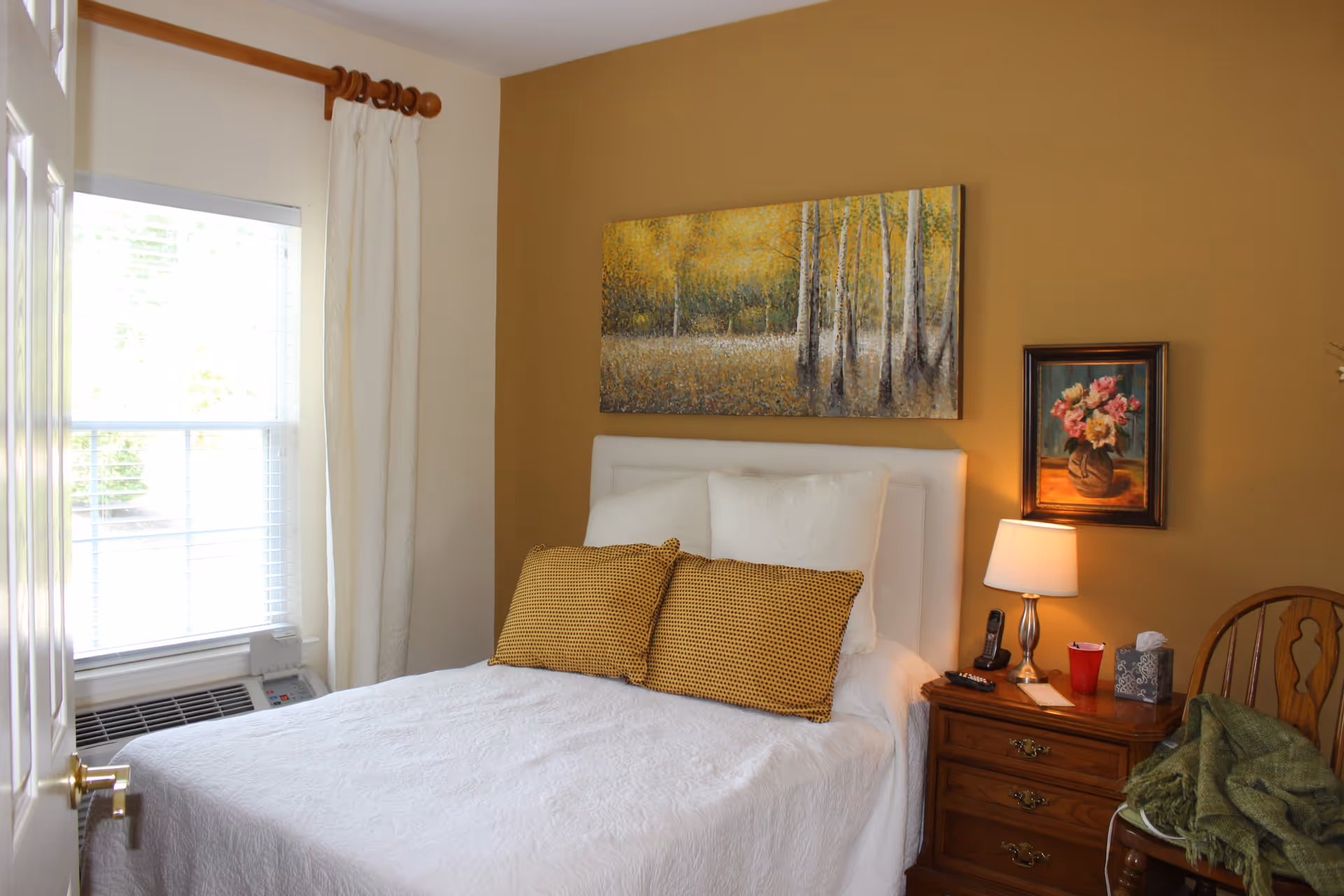 A cozy bedroom with a white bedspread and two yellow patterned pillows on a white headboard. The room has a mustard yellow accent wall with two paintings, one large landscape and one smaller floral. A wooden nightstand with a lamp, phone, remote control, red cup, and tissue box is beside the bed. A wooden chair with a green blanket is next to the nightstand. A window with white curtains and blinds lets in natural light.