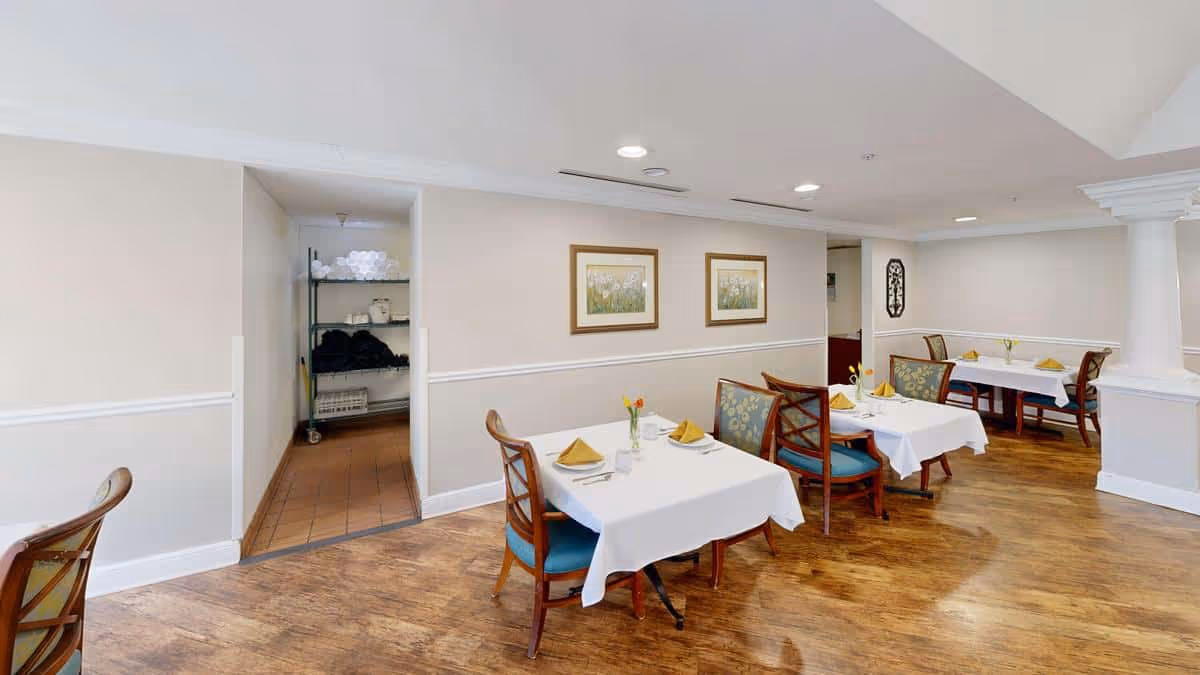 A dining room in Pegasus Landing of Overland Park featuring several tables with white tablecloths, each set with yellow napkins and small flower vases. The room has wooden floors, beige walls with white trim, and framed floral artwork on the walls. There is an open doorway leading to a storage area with shelves holding towels and other supplies.
