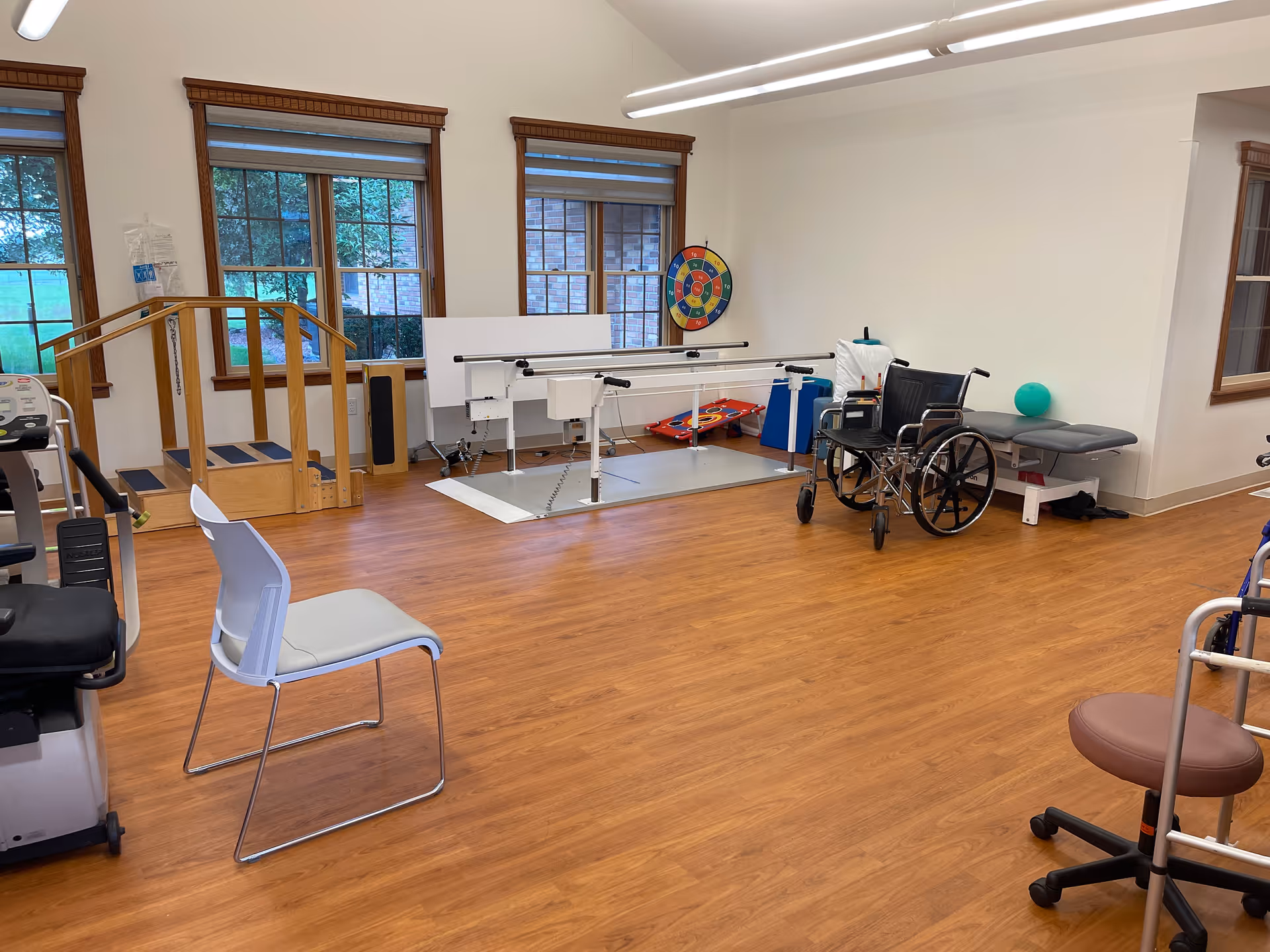 Physical therapy/rehab room with parallel bars, a wheelchair, chairs, and exercise equipment on a wood floor.