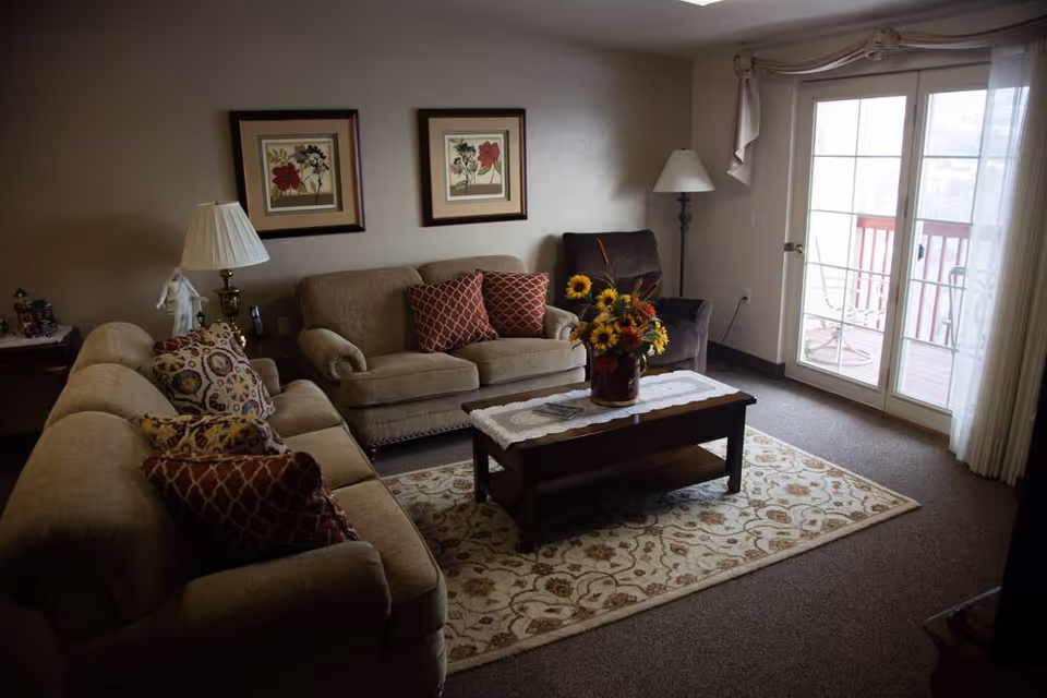 Cozy living room with two sofas, an armchair, a coffee table topped with a floral arrangement, framed art on the wall, and sliding glass doors to a balcony.