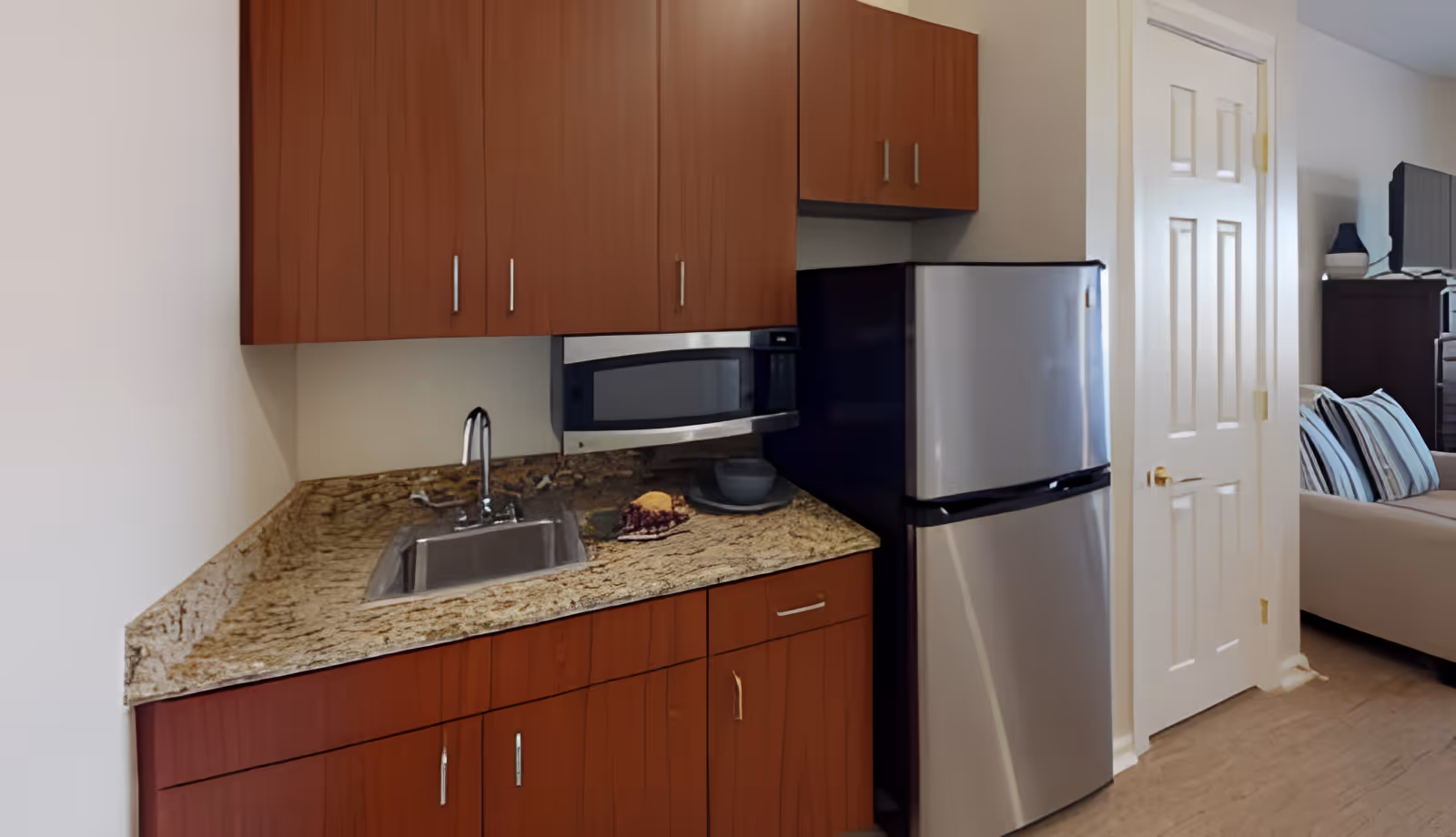 A small kitchen area with wooden cabinets, a granite countertop, a stainless steel sink, a microwave, and a stainless steel refrigerator. To the right, there is a white door and part of a living room with a beige couch and striped pillows is visible.