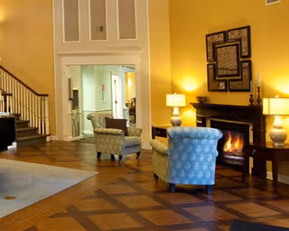 A cozy living room area with two patterned armchairs facing a lit fireplace. The room has warm yellow walls, wooden flooring with a geometric pattern, two table lamps on either side of the fireplace, and decorative artwork above the mantel. A staircase is visible on the left side, and an open doorway leads to another room in the background.