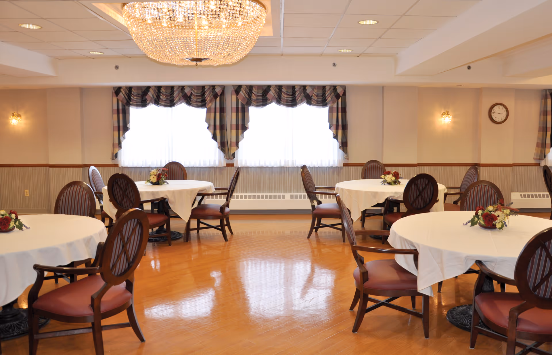 A dining room with round tables covered in white tablecloths, each table decorated with a small floral centerpiece. Wooden chairs with cushioned seats surround the tables. The room has polished wooden floors, large windows with plaid valances, wall sconces, and a large chandelier hanging from the ceiling.