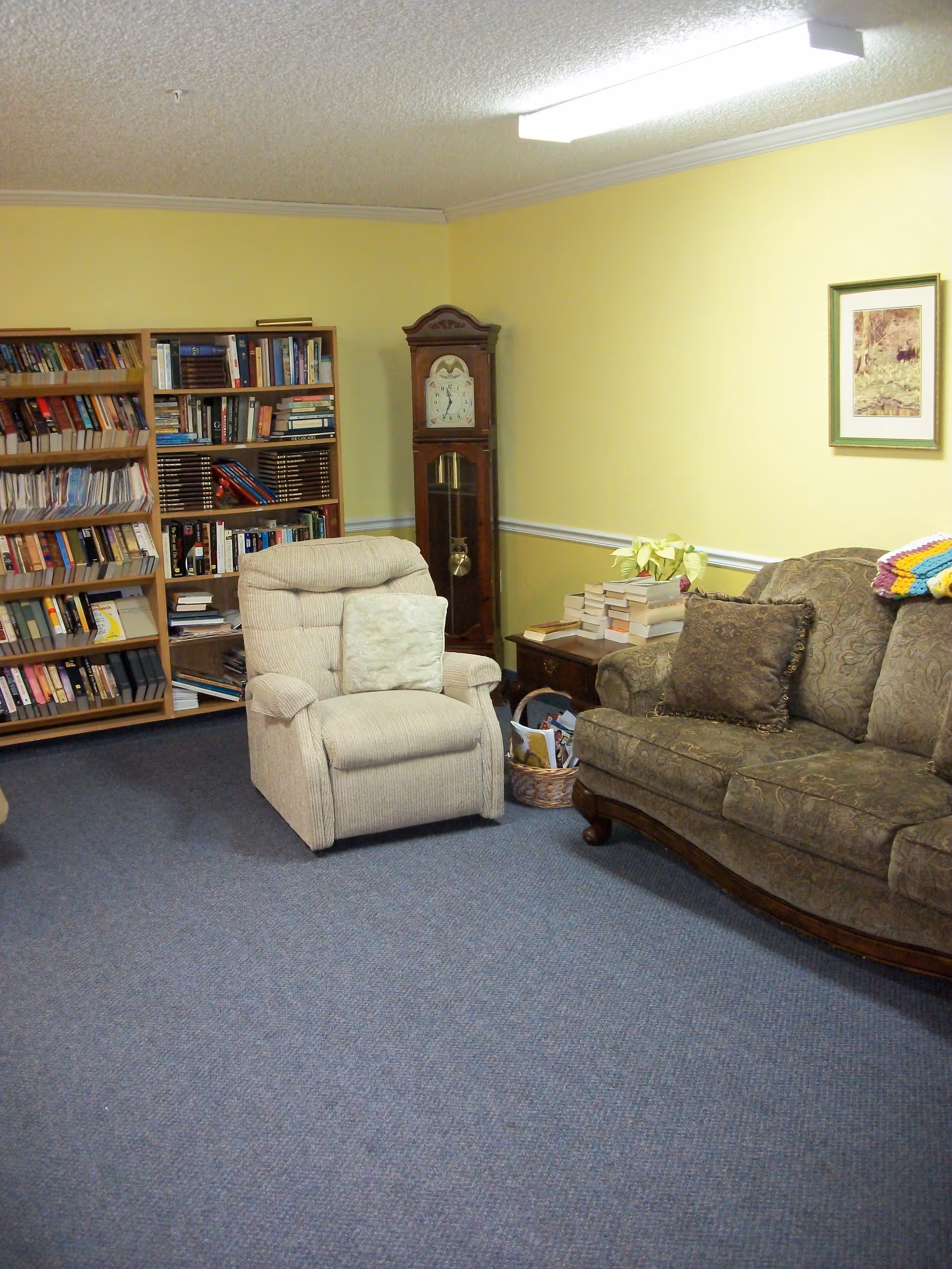 A cozy lounge with bookshelves, a recliner and sofa, a grandfather clock and a side table piled with books.