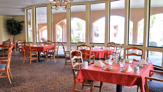Dining room with several wooden tables and chairs, each table covered with a red tablecloth and set with plates, glasses, napkins, and silverware. Large windows line the wall, letting in natural light and showing an outdoor covered walkway with arches.
