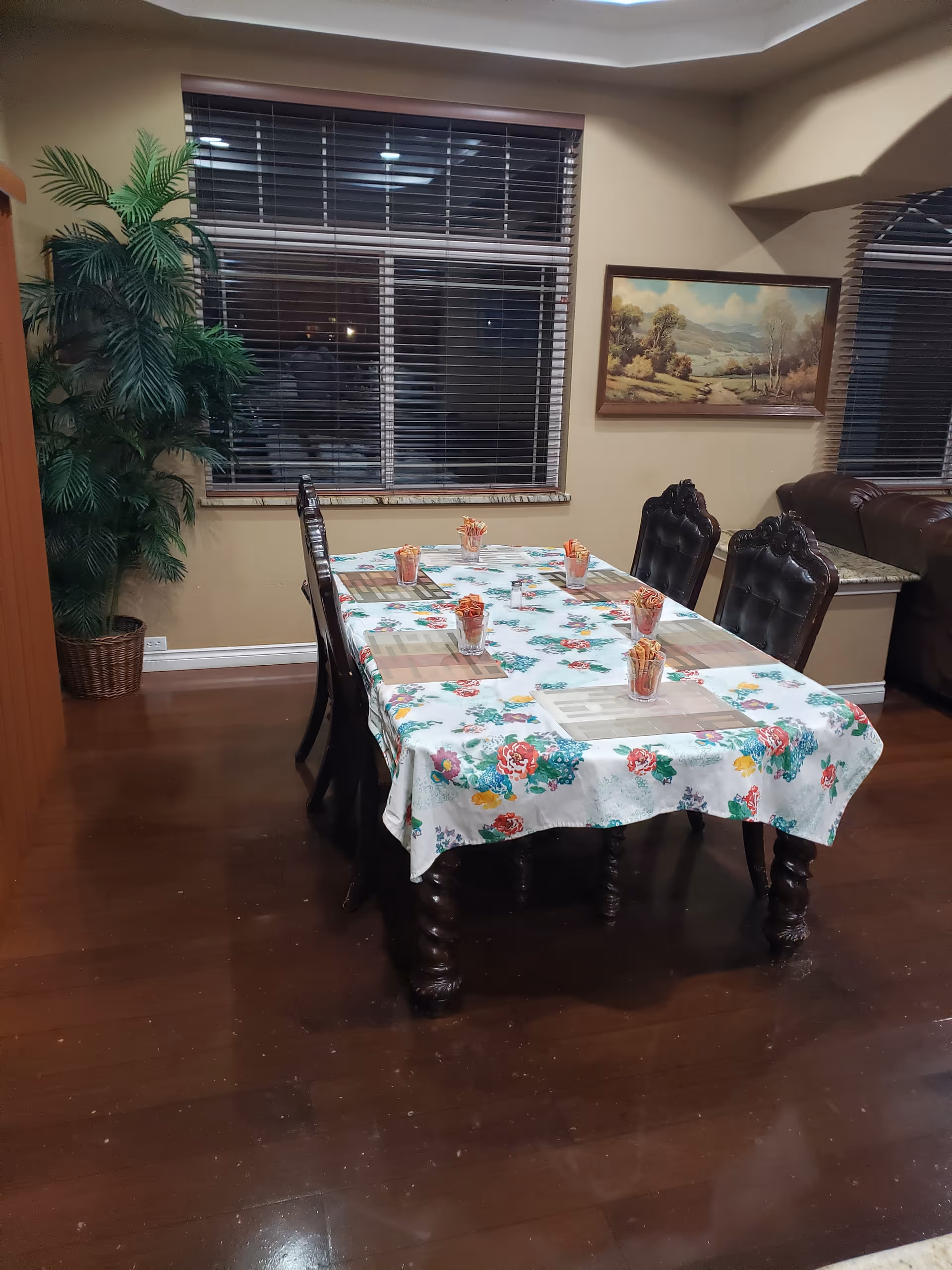 Dining room with a rectangular wooden table covered with a floral tablecloth. Six dark wooden chairs with ornate backs surround the table. Each place setting has a placemat and a glass filled with wrapped candies. There is a large window with blinds behind the table, a potted plant in the corner, and a landscape painting on the wall. The floor is dark wood.