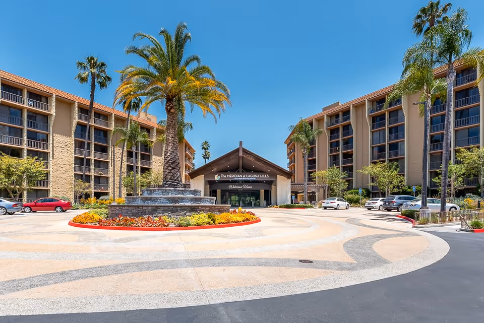 Front exterior view of Meridian at Laguna Hills facility with a circular driveway, a central landscaped area featuring a tall palm tree and colorful flowers, flanked by two multi-story buildings under a clear blue sky.