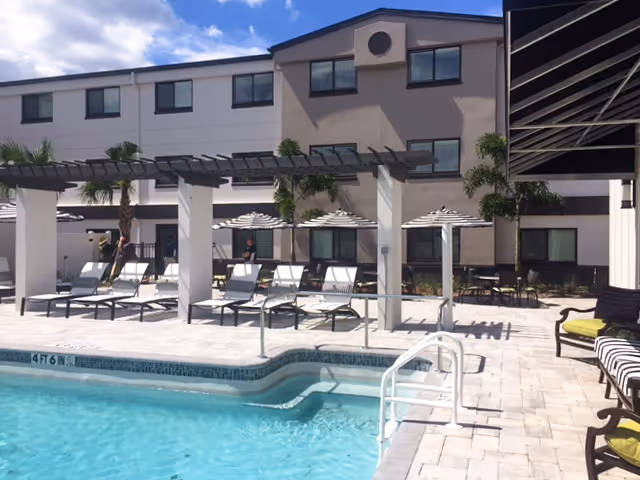 Outdoor swimming pool area with lounge chairs and umbrellas next to a three-story building under a partly cloudy sky. There are palm trees and shaded seating areas around the pool.