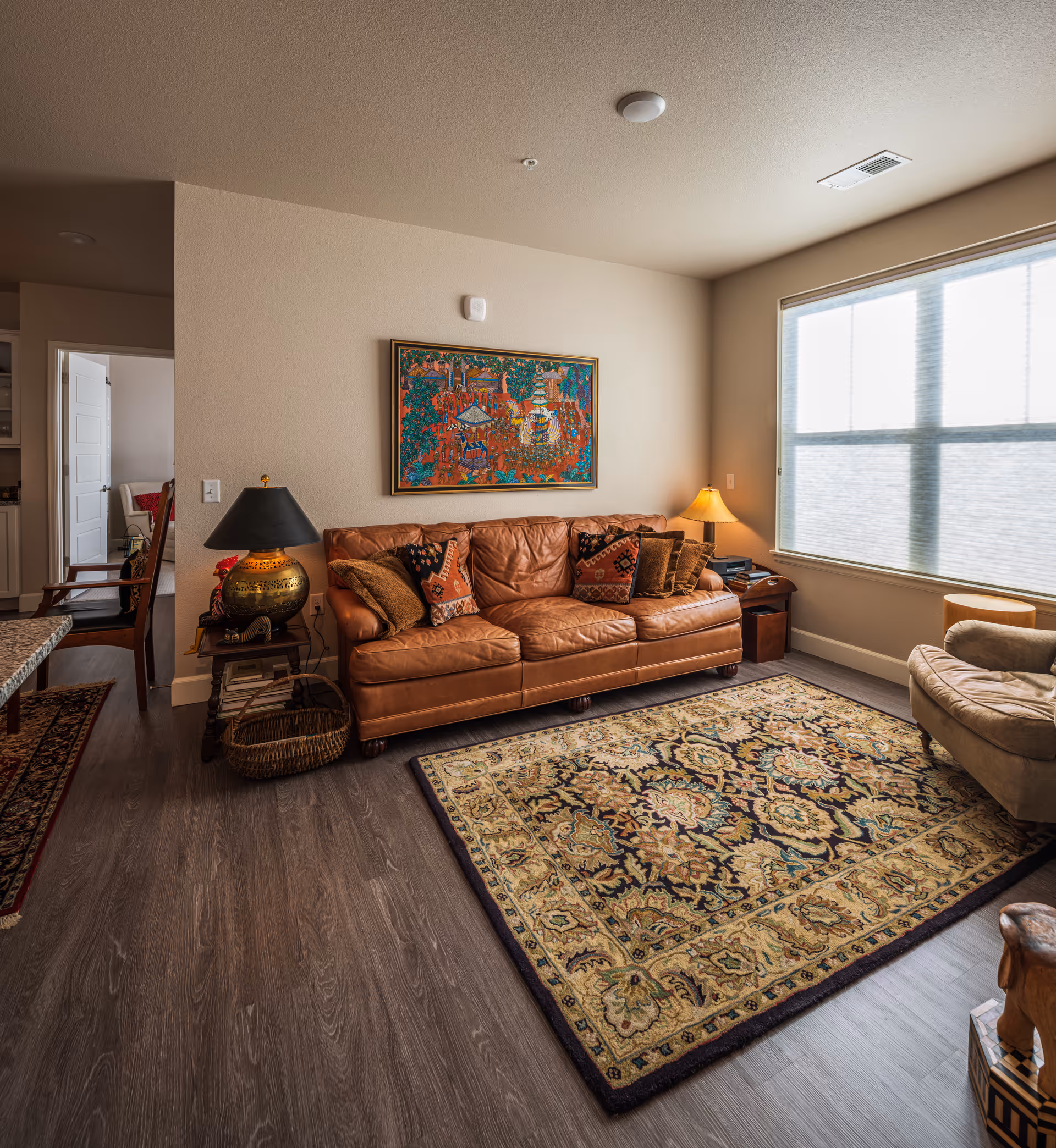 A cozy living room with a brown leather sofa adorned with patterned cushions, a decorative area rug with intricate designs, a side table with a lamp, and a large window with blinds allowing natural light to fill the room. A colorful framed painting hangs on the wall above the sofa. The room has wooden flooring and neutral-colored walls.