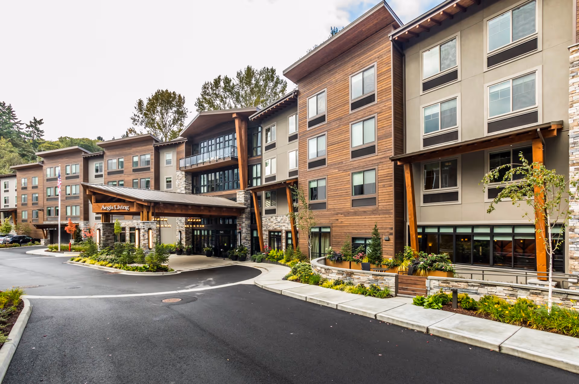 Exterior view of Aegis Living Mercer Island, a multi-story senior living facility with a combination of wood and stone facade, large windows, and a covered entrance. The surrounding area includes landscaped greenery and a paved driveway.