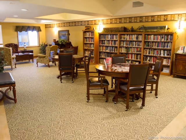 A cozy senior living library/common room with bookshelves, round tables and chairs, and lounge seating.
