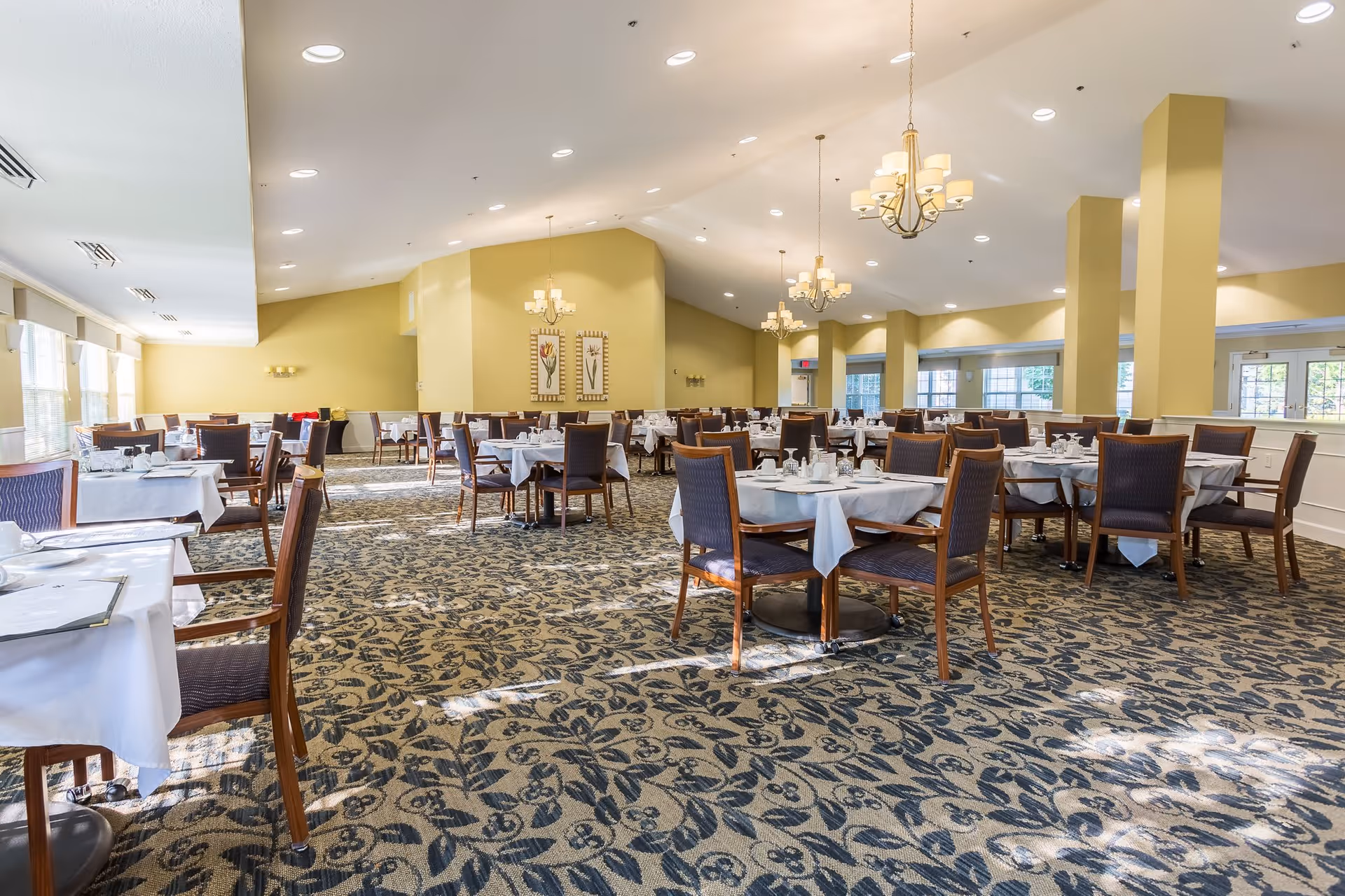 A spacious dining room with multiple tables covered in white tablecloths, each set with plates, cups, and silverware. The room has patterned carpet flooring, yellow walls, large windows letting in natural light, and several chandeliers hanging from the ceiling.