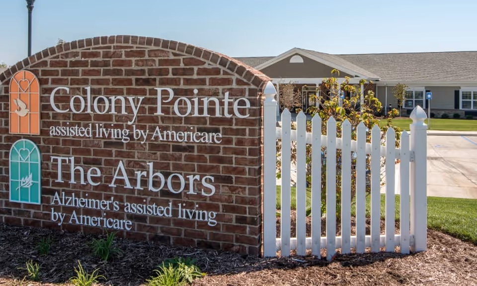A brick sign at the entrance of Colony Pointe Senior Living and The Arbors Alzheimer's assisted living by Americare, next to a white picket fence with a building and parking area in the background under a clear blue sky.