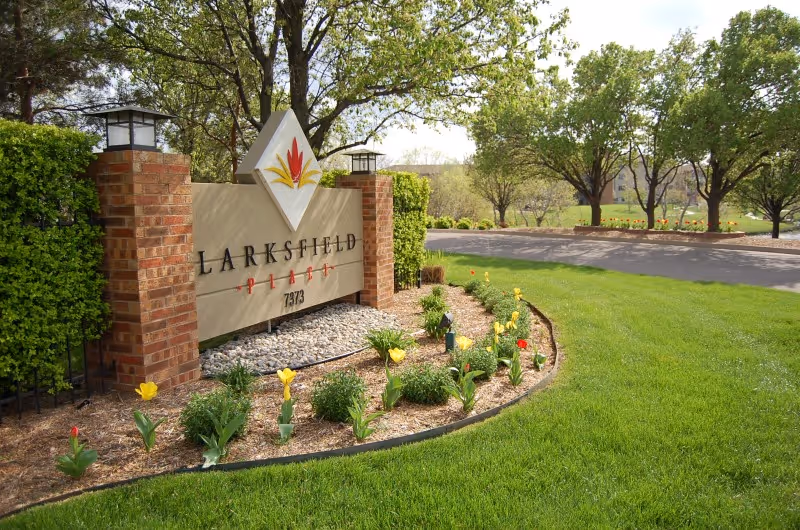 Entrance sign for Larksfield Place Independent Living surrounded by a landscaped garden with yellow and red tulips, green grass, and trees in the background under a clear sky.