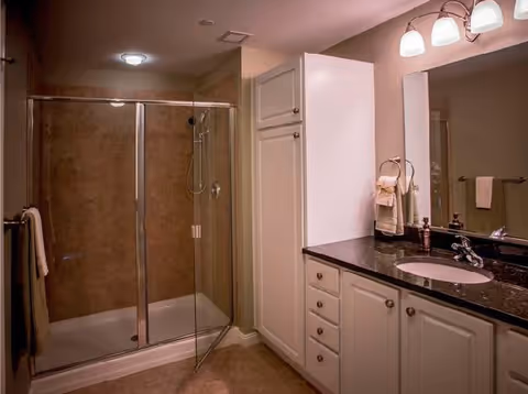 A bathroom with a glass-enclosed shower on the left, beige tiled walls inside the shower, a white cabinet with drawers and doors, a black countertop with a sink, a mirror above the sink, and a light fixture with three bulbs above the mirror. Towels hang on a ring and a bar near the shower.