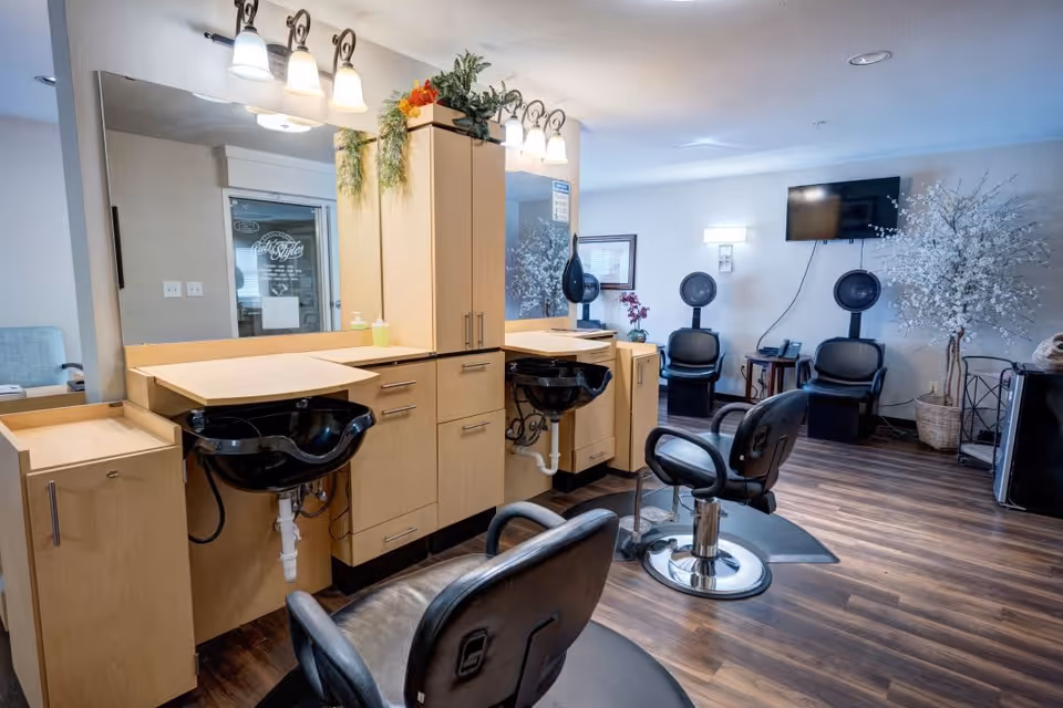 Interior view of a hair salon area in a senior living facility with two black salon chairs in front of wooden cabinets and sinks. The room has wooden flooring, a large mirror, and hair drying chairs with hooded dryers in the background. There is a TV mounted on the wall and decorative plants in the corner.