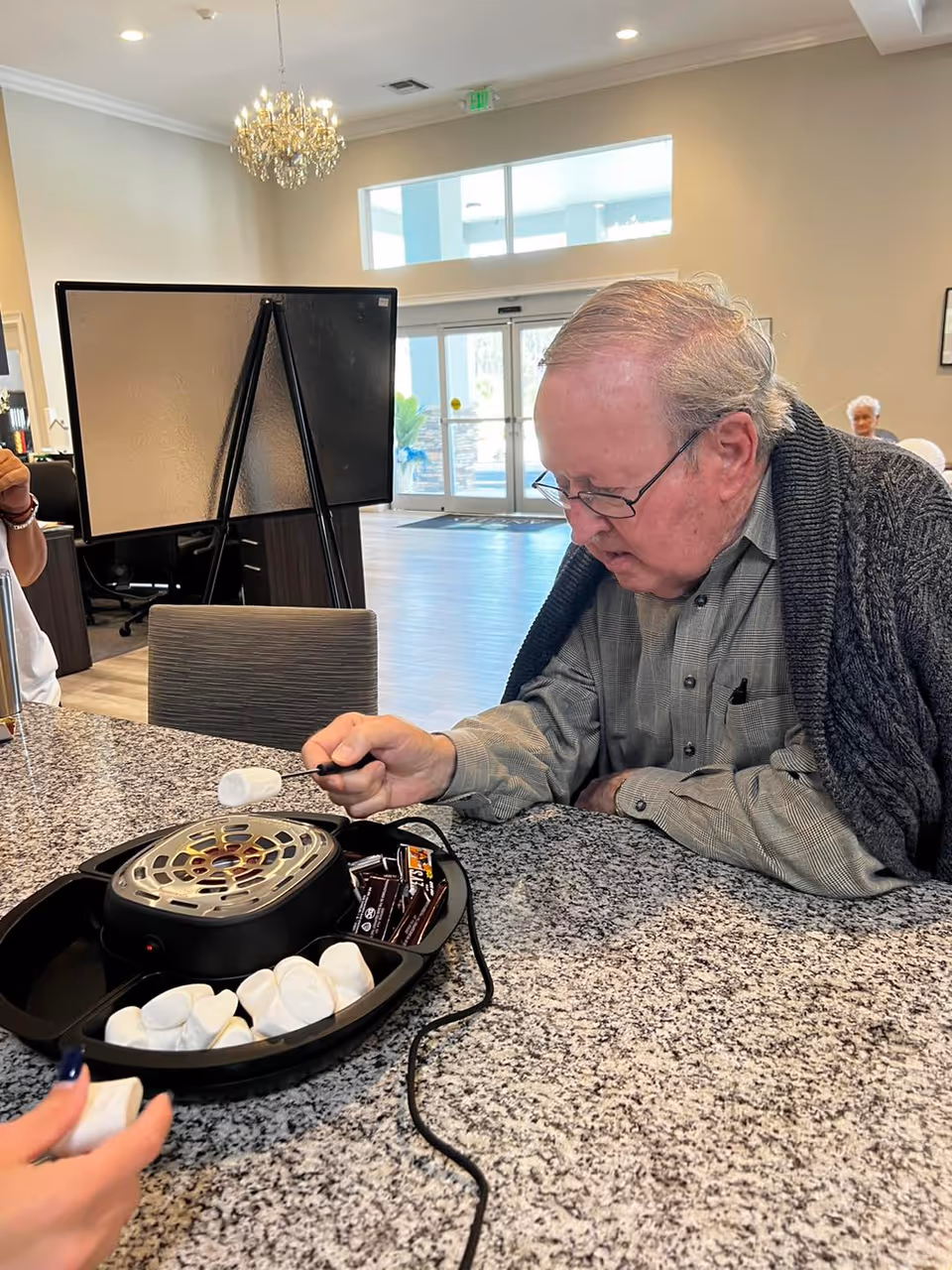 An elderly man with glasses and a gray sweater is roasting a marshmallow over an electric s'mores maker on a granite countertop in a bright, spacious room with large windows and a chandelier. Another person’s hand is visible holding a marshmallow nearby.