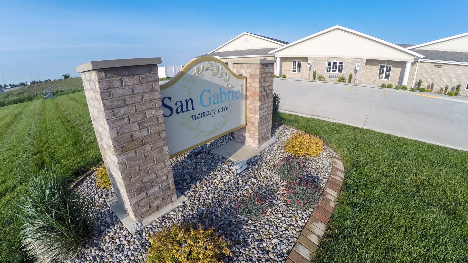 Outdoor view of the San Gabriel Memory Care facility sign mounted between two brick pillars surrounded by landscaping with rocks, plants, and grass. The building and parking lot are visible in the background under a clear blue sky.