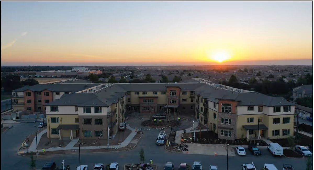 Aerial view of a large three-story senior living facility named Bruceville Point at sunset, showing the building's U-shaped layout with parking spaces and surrounding neighborhood in the background.