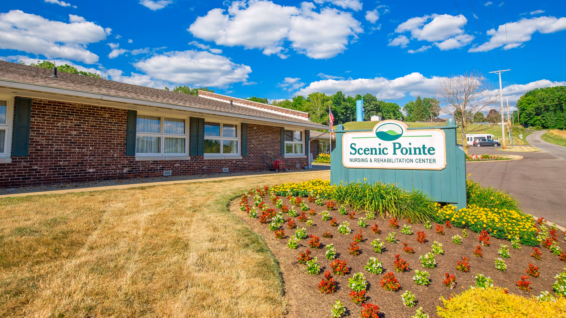 Exterior view of Scenic Pointe Nursing & Rehabilitation Center showing a brick building with windows, a well-maintained flower bed with colorful flowers, a green sign with the facility name, and a partly cloudy blue sky.