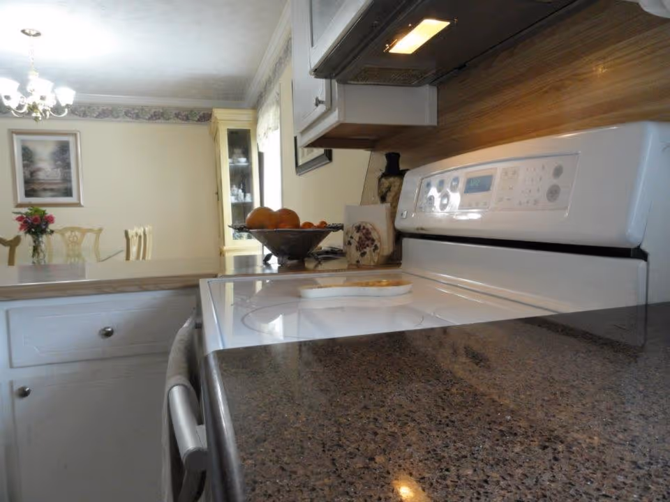 View of a kitchen area featuring a white electric stove with a digital display, a dark speckled countertop, and white cabinetry. In the background, there is a dining area with a table, chairs, a chandelier, a framed picture on the wall, and a glass-front cabinet. A bowl of fruit and a vase with flowers are also visible on the counter and table respectively.