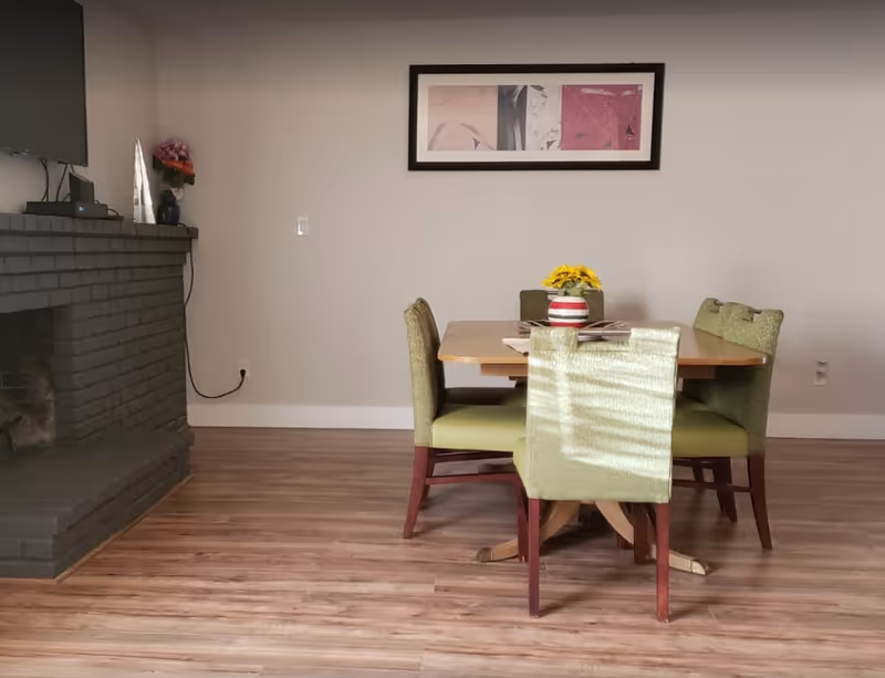 Small dining area with a wooden table and four green chairs, a striped vase with flowers on the table and a gray brick fireplace to the left.