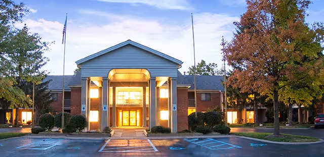 Front entrance of a two-story brick retirement living building with a lit portico and columns, parking spaces, and trees.