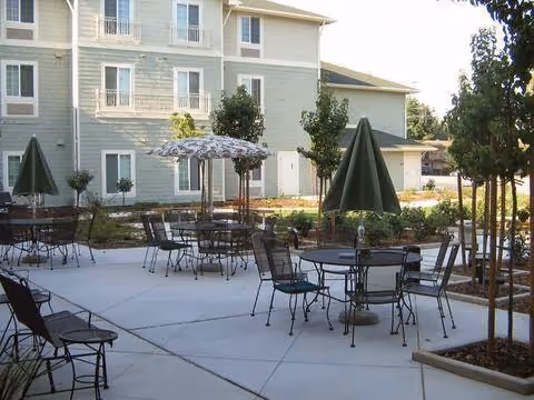 Outdoor courtyard with metal patio tables, chairs, and umbrellas in front of a multi-story residential building.