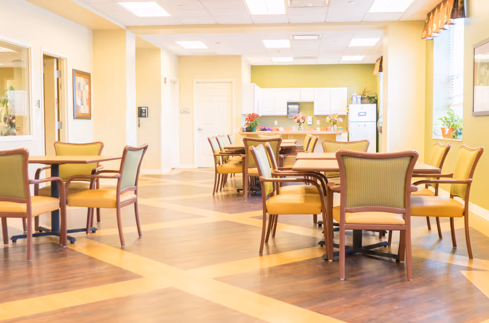 A bright dining area in a senior living facility with multiple wooden tables and chairs arranged neatly. The room has light yellow walls, large windows with valances, and a kitchen area with white cabinets and a refrigerator in the background. The floor has a patterned wood design.