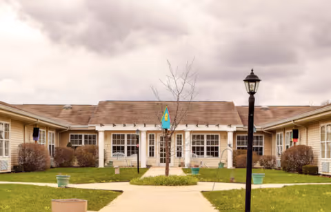 Courtyard with a central walkway, grass, planters and a lamppost leading to a single-story building with multiple windows under a cloudy sky.