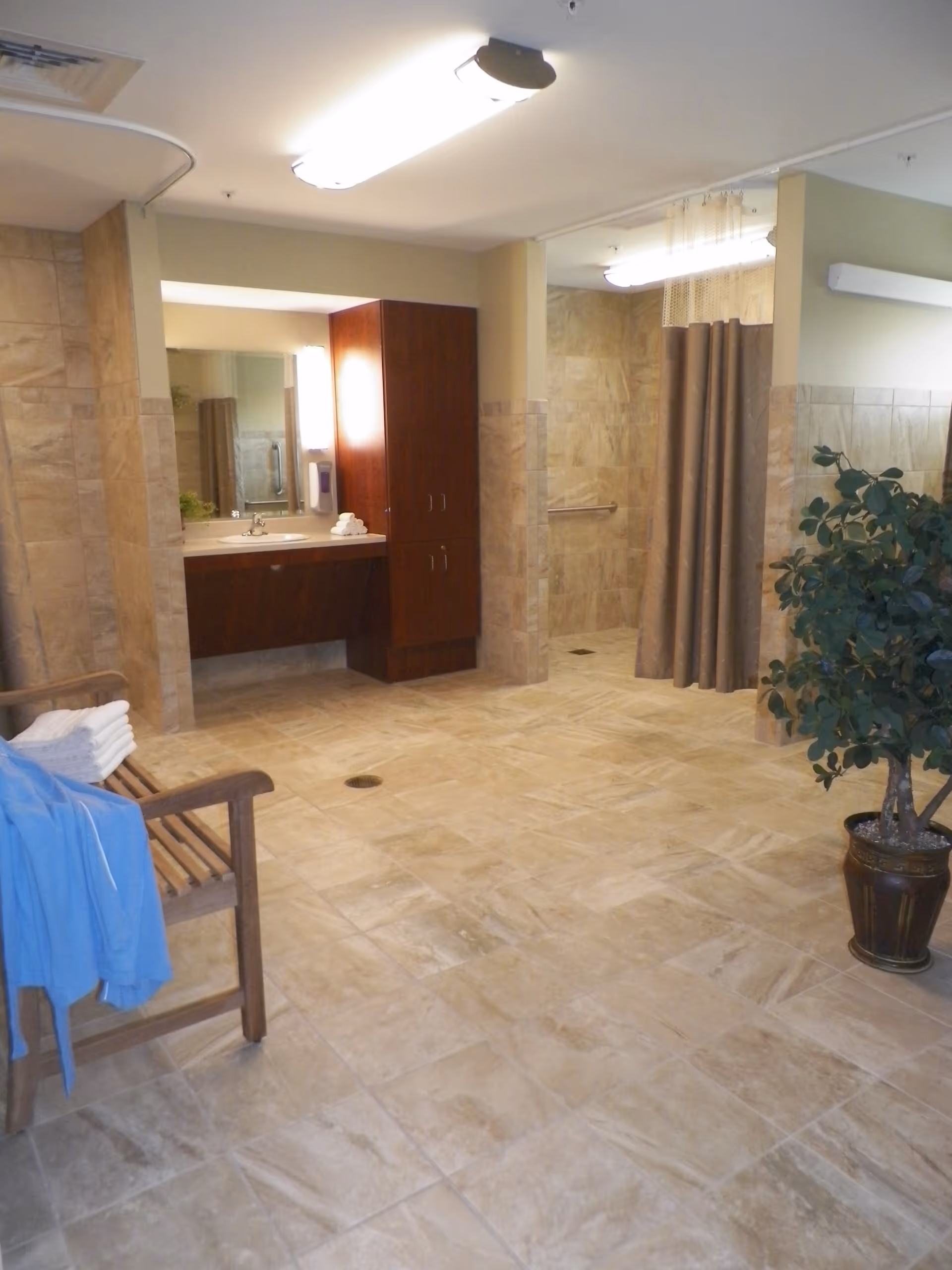 Spacious tiled accessible bathroom with a vanity and mirror, curtained shower stalls, a wooden bench with towels, and a potted plant.