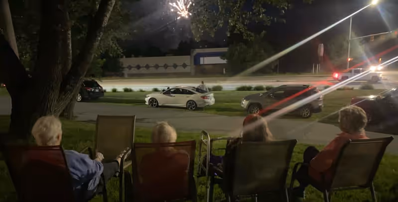Four elderly people sitting on chairs outside at night watching fireworks in the sky above a street with parked cars and trees nearby.