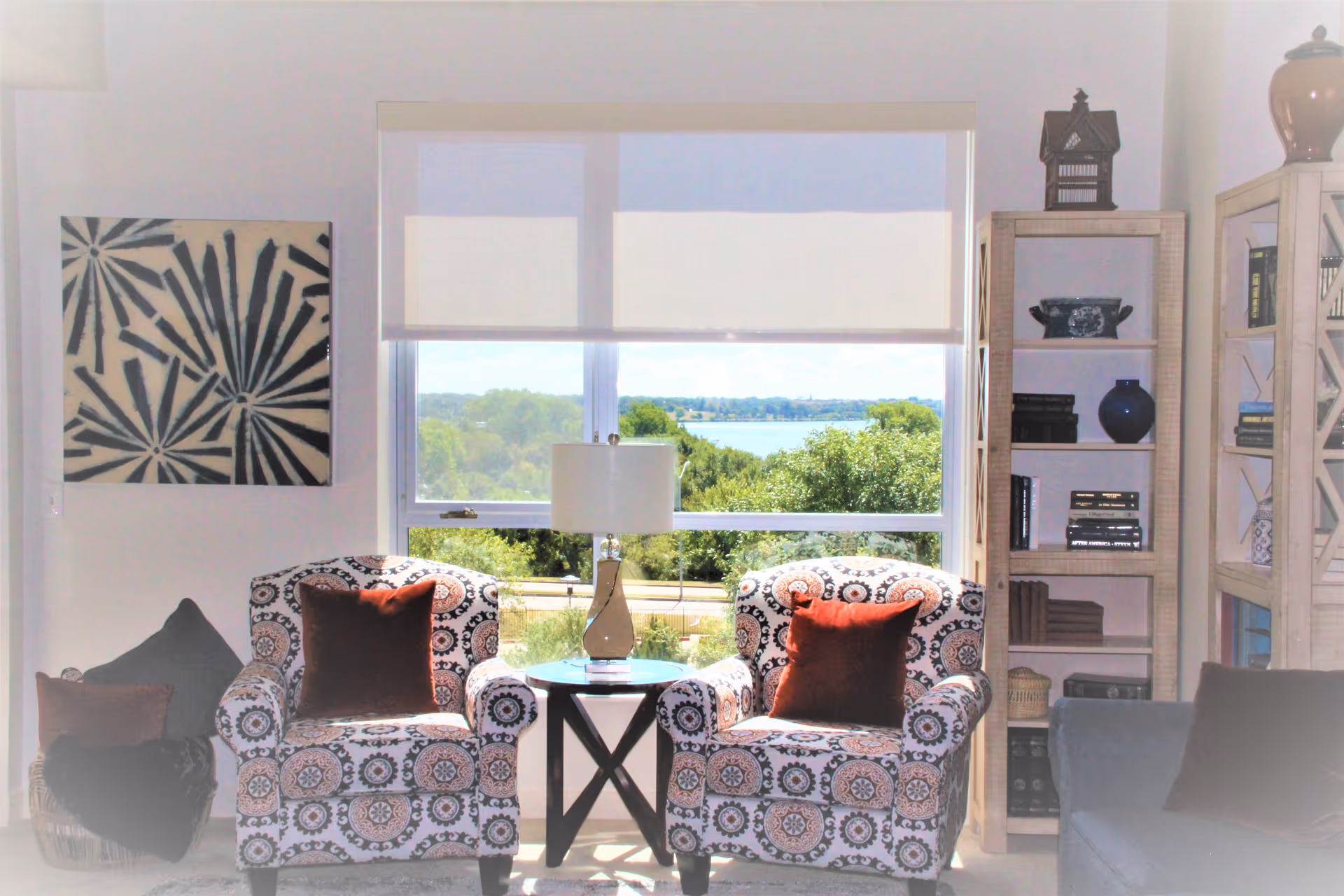 A bright living room area with two patterned armchairs each with a rust-colored pillow, a small round table with a lamp between them, a large window with a view of trees and water outside, a bookshelf filled with books and decorative items on the right, and a piece of abstract wall art on the left.