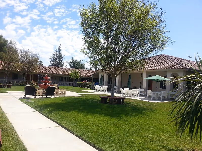 Outdoor courtyard area of a senior living facility with green grass, a tree with a circular bench around it, several white tables and chairs with green umbrellas, a red multi-tiered fountain, and a building with a tiled roof in the background under a partly cloudy sky.