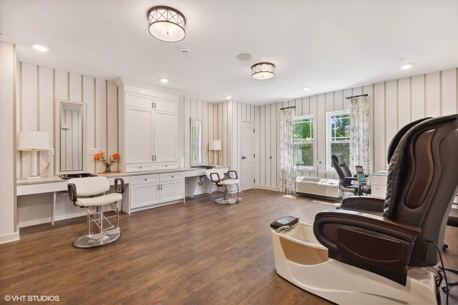 A bright and spacious salon room with wooden flooring, two white styling chairs in front of mirrors on the left wall, and two large black pedicure chairs with foot baths on the right side. The room has white walls with vertical stripes, two windows with floral curtains, and ceiling lights.