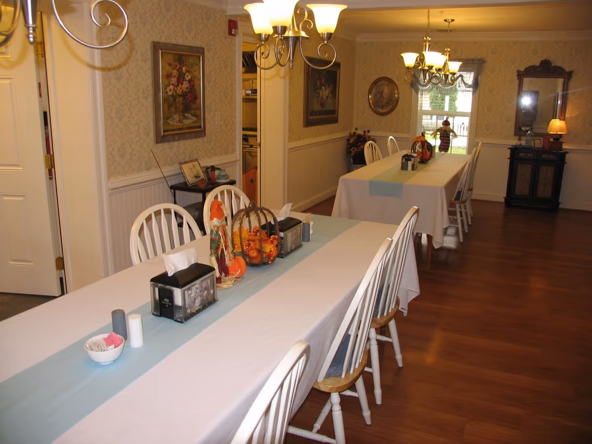 A dining room with two long tables covered with white tablecloths and light blue runners. Each table is surrounded by white wooden chairs. The tables have decorative items including a scarecrow figure, a basket with small pumpkins, napkin holders, and condiments. The room has wooden flooring, wallpapered walls with floral paintings, a mirror, and two chandeliers hanging from the ceiling. A window at the far end shows a person outside.