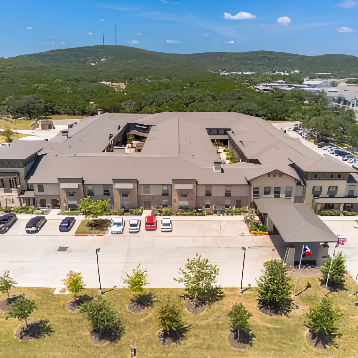 Aerial view of a large senior living facility building with a beige roof and multiple windows, surrounded by a parking lot with several cars and landscaped greenery. In the background, there are green hills under a blue sky with a few clouds.