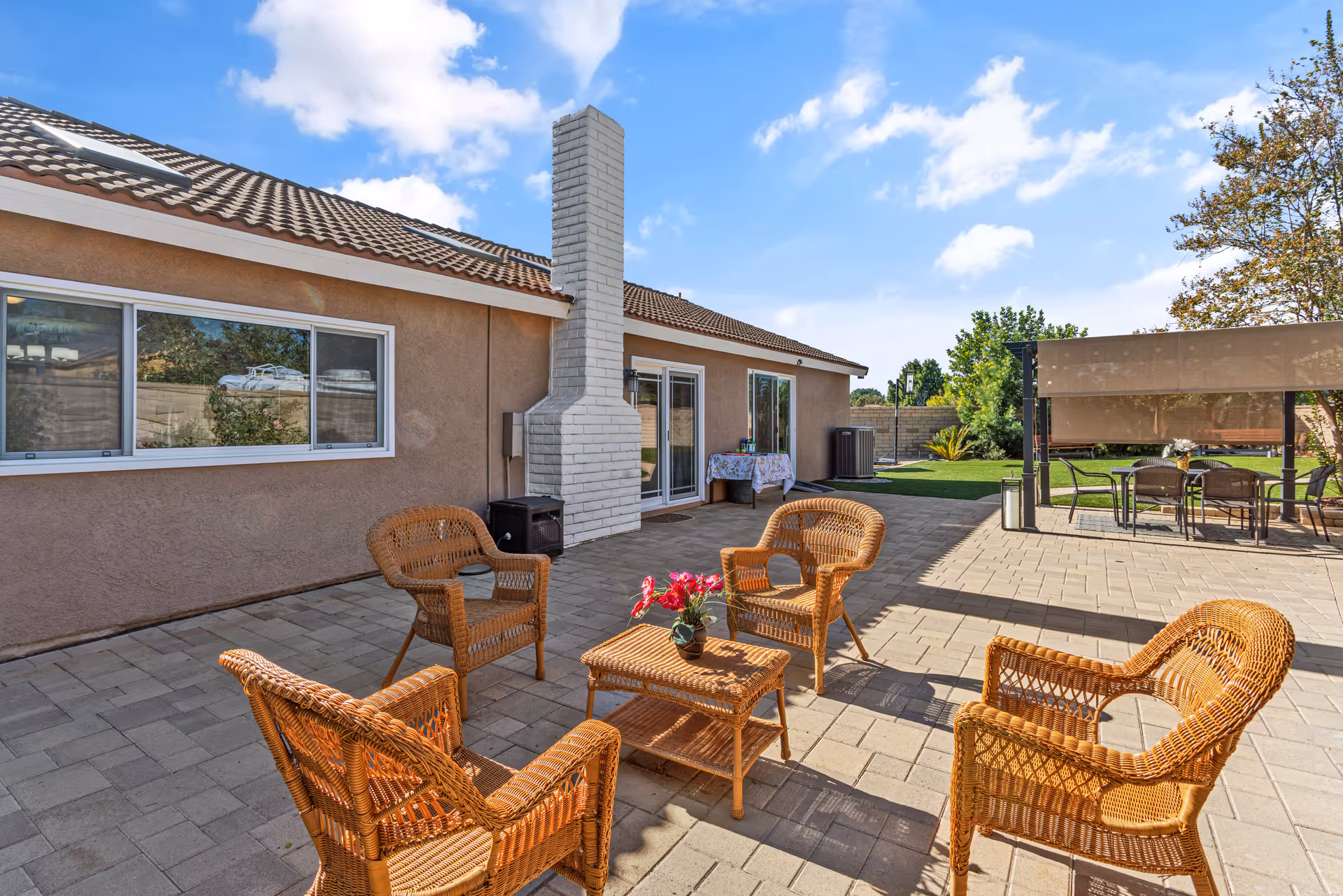 Outdoor patio area with four wicker chairs arranged around a small wicker table with a flower pot on it. The patio is paved with stone tiles and is adjacent to a beige building with a white brick chimney. In the background, there is a shaded seating area with a table and chairs, green grass, trees, and a clear blue sky with some clouds.