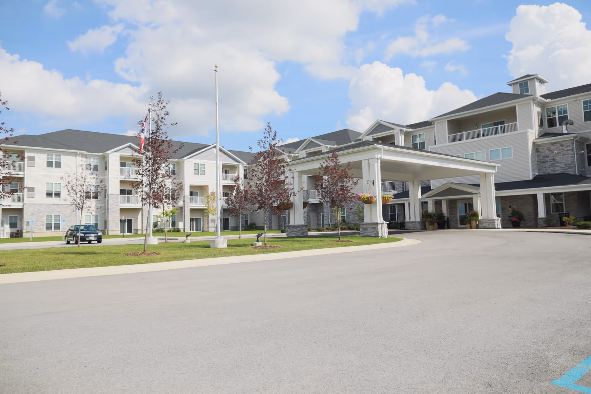 Exterior view of StoryPoint Waterville senior living facility showing a large multi-story building with balconies, a covered entrance, a driveway, small trees, and a flagpole under a partly cloudy sky.